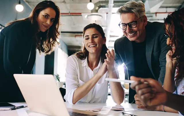 Four professionals smiling and engaging in a discussion around a laptop in a modern office setting.