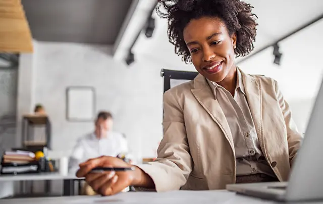 A woman in a beige blazer smiles while writing notes, with a laptop in front of her. 