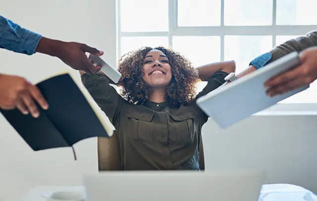 A smiling woman sitting at a desk with headphones, relaxing with hands behind her head while receiving books from others. 