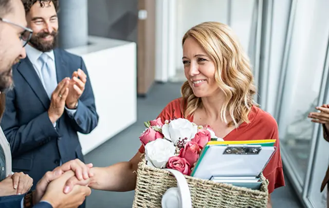 A blonde woman in a reddish-orange top smiles warmly as she shakes hands with a male colleague in a modern office hallway.