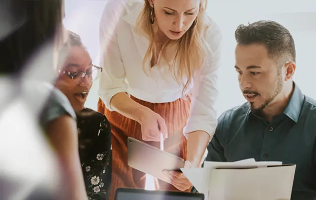 A group of three people engaged in a discussion, one showing a tablet to others. 