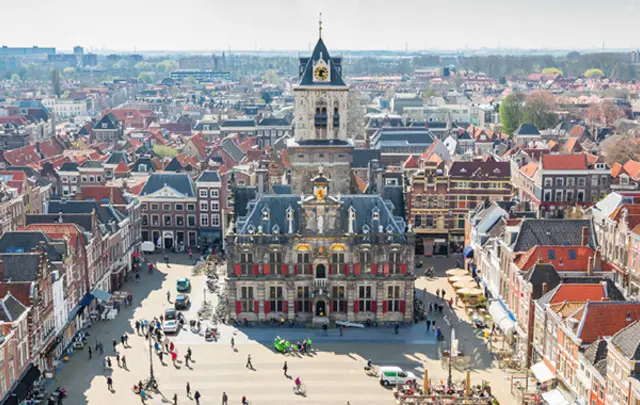 Aerial view of a historic town square with a large clock tower and red-roofed buildings. 