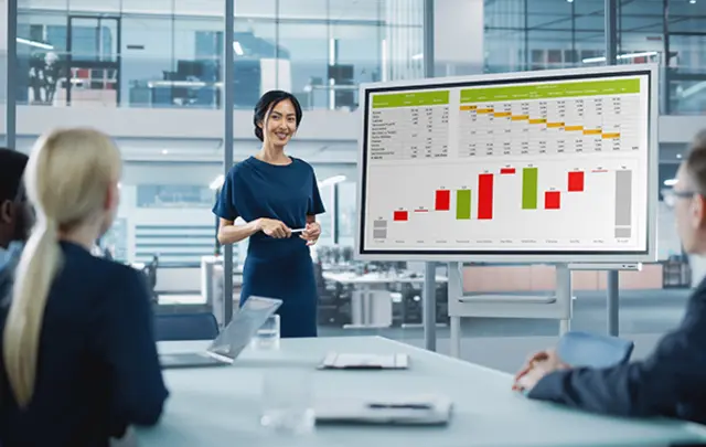 A woman presents data in front of a screen displaying colorful graphs to four listeners in an office. 