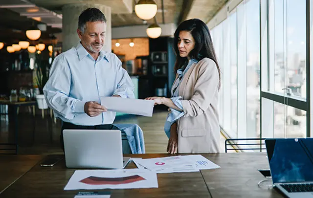 Two professionals discussing documents at a table with laptops and papers, in a modern office. 