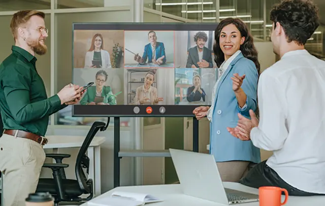 A group of six people in a modern office meeting, discussing at a large table with laptops and mugs. 