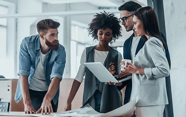 Four people in a modern office discussing a project on a laptop and paperwork. 