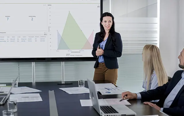A woman presents data on a screen to a meeting room of people with laptops and papers. 