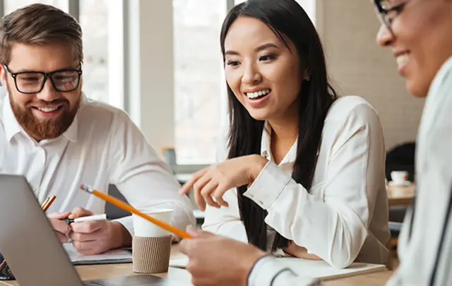 Three people collaborating at a table with a laptop, notebook, and coffee. 