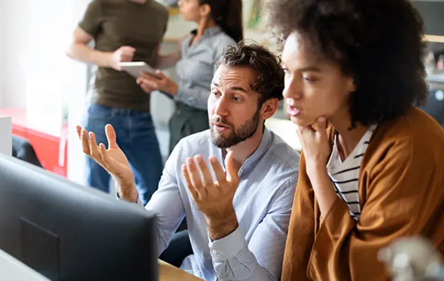 A man and woman discussing something on a computer screen in a busy office. 