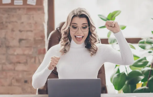 A happy woman in a white sweater celebrates while looking at her laptop. 