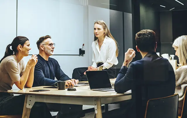 A team meeting in a modern office with six professionals at a table, discussing with a laptop and coffee cups. 