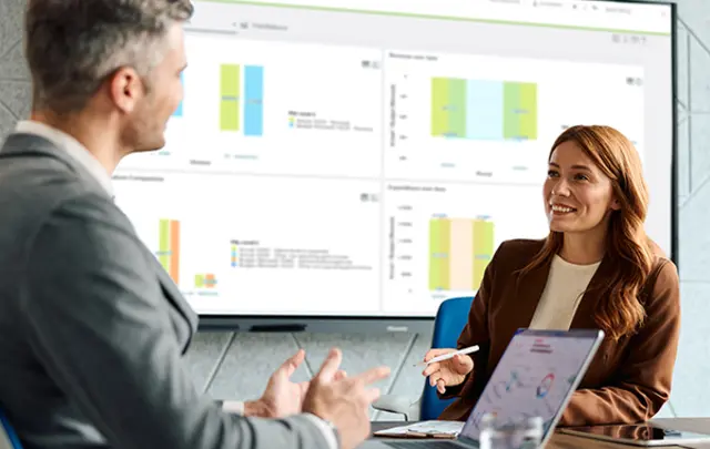 A woman and a man in a meeting discussing charts and data displayed on a screen. 