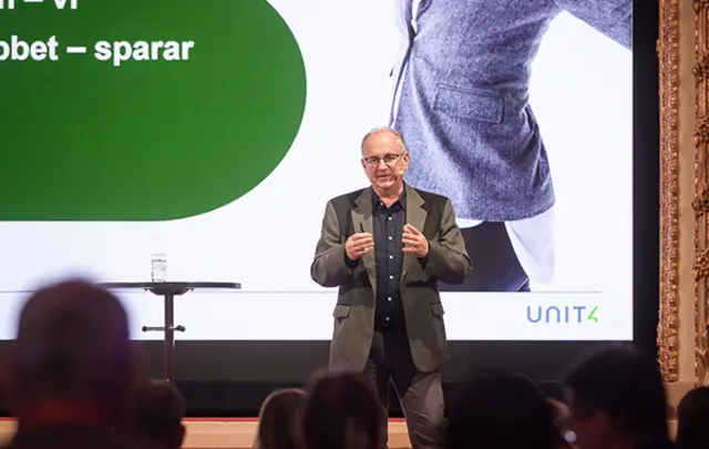 A speaker in a gray suit presents on stage in front of a large screen. 
