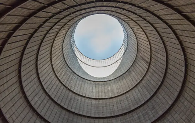 View looking upward through a series of concentric circular concrete rings forming an industrial structure, with an open circular skylight at the top revealing a patch of blue sky. 