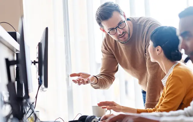 A person standing beside a seated colleague in an office setting, gesturing toward a computer screen while discussing work. 