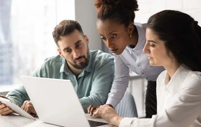 Three people working together at a laptop in an office, focused on the screen. 
