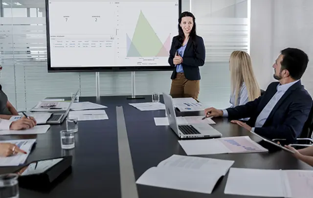 A group of professionals sit around a conference table during a meeting while a presenter stands at the front, gesturing toward a large screen displaying charts and graphs. 