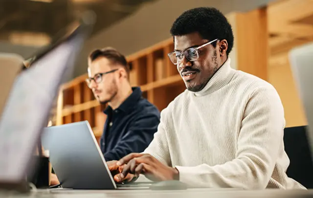 Several people are seated at a long desk in a modern, warm‑toned workspace, working on laptop computers. 