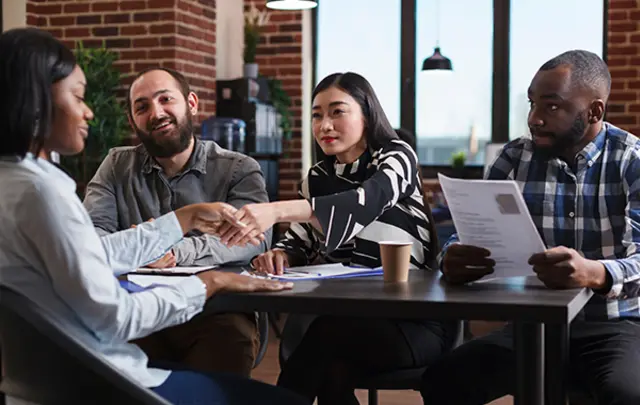 A diverse group of professionals seated around a table in a modern office, reviewing printed documents and exchanging papers during a collaborative meeting, with brick walls, large windows, and pendant lighting in the background.