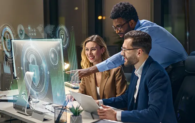 A group of three people collaborate in a modern office at night, working around a large transparent digital display showing futuristic security‑themed graphics, including a padlock icon and circular data visualizations.