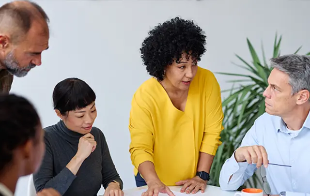 A group of five people gathered around a table in a bright meeting room, reviewing documents and discussing work. 