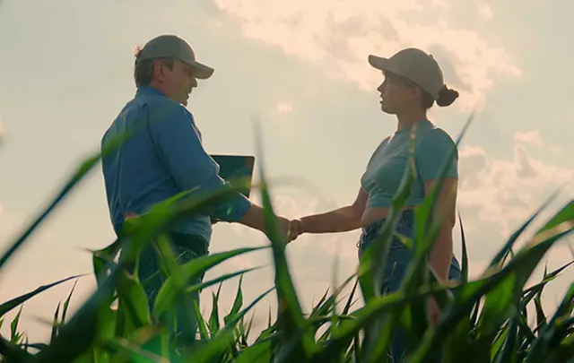 wo individuals standing in a tall crop field shaking hands, with one holding a laptop. 