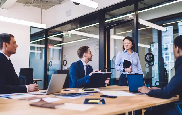 A group of professionals collaborating in a modern office meeting room, seated around a table with laptops and documents, while one colleague stands and presents ideas near glass walls and large windows.