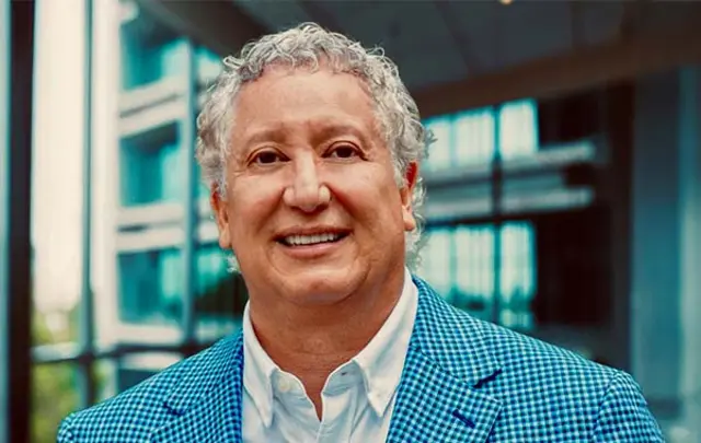 Business professional wearing a blue checkered blazer and white shirt, standing indoors in a modern office environment with glass walls and natural light in the background.
