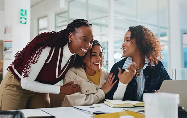 Three people collaborate at a table in a bright, modern learning space, discussing notes and ideas while working with a laptop, notebooks, and documents, suggesting teamwork and academic or professional collaboration.