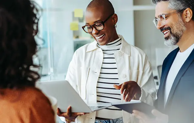 Three professionals stand in a modern office environment, reviewing information on a tablet together while discussing a document, suggesting collaboration and decision‑making in a workplace setting.