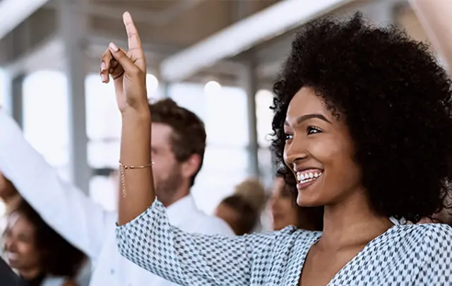 A smiling woman raises her hand enthusiastically during a business meeting or training session, with colleagues doing the same in the background.