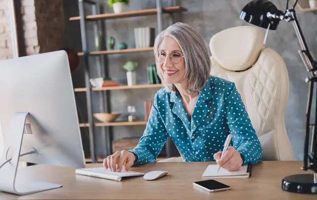 A professional woman sits at a desk in a modern home office, working on a desktop computer while taking notes in a notebook, with shelves, plants, and a desk lamp visible in the background.