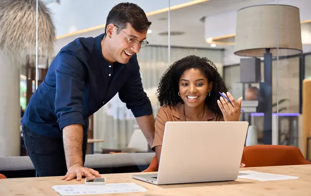 Two professionals collaborating at a desk in a modern office, reviewing documents and discussing insights while working on a laptop in a bright, contemporary workspace.