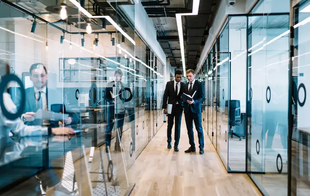 Two business professionals walking and discussing documents in a modern office corridor with glass meeting rooms, workspaces visible on both sides, and contemporary lighting.
