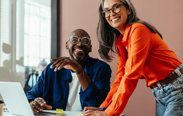 Two colleagues working at a desk with a laptop, reviewing documents together in a modern office environment.