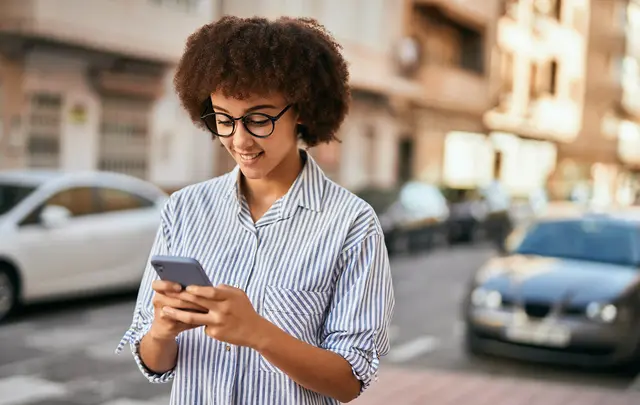 Person standing on a city street, wearing a striped button-down shirt and holding a smartphone, with parked cars and residential buildings in the background.