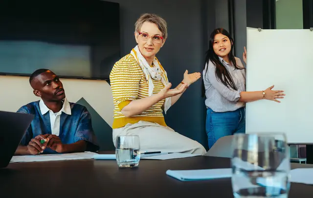 A group of people in a meeting room reviewing information on a whiteboard and documents during a collaborative procurement or contract management workshop.