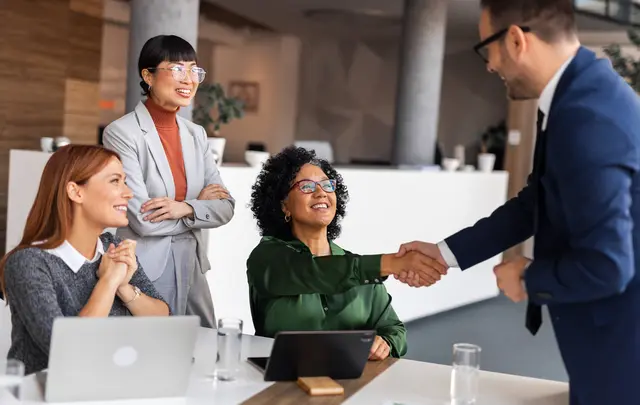 Business professionals collaborating at a meeting table, with one person shaking hands while others observe, laptops and tablets visible in a modern office setting.