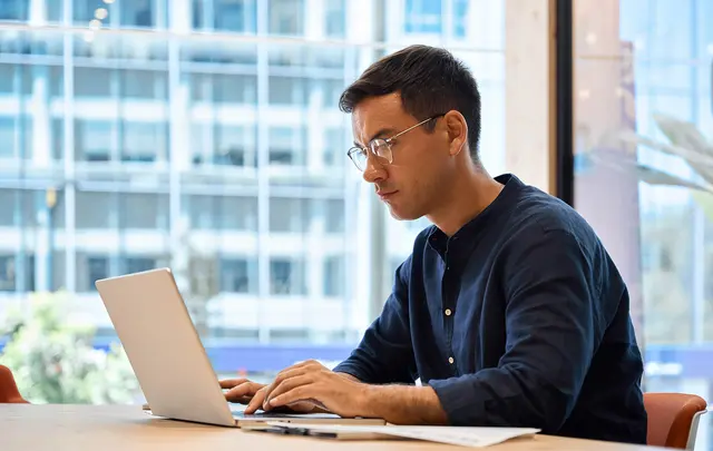 Person working on a laptop at a desk near a large window, with modern office buildings visible outside.