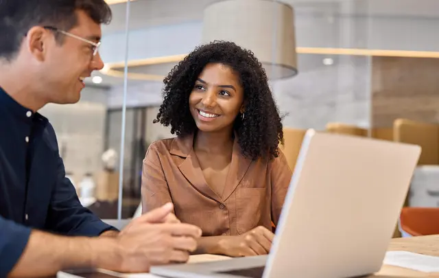 Two colleagues seated at a table in a modern office, collaborating while reviewing information on a laptop during a focused discussion.
