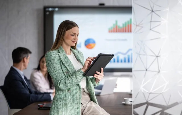 A professional using a tablet during a finance planning meeting, with data charts displayed on a screen in the background.