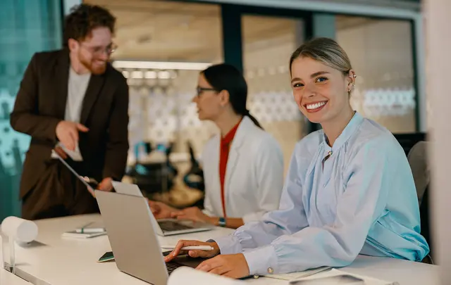 Three people working at laptops in a modern office, with one person in the foreground seated at a desk and two colleagues collaborating in the background.