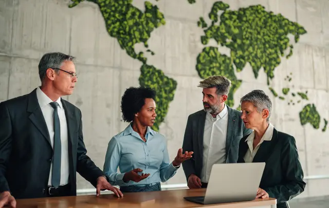 Four professionals stand around a table with a laptop, discussing work in a modern office.