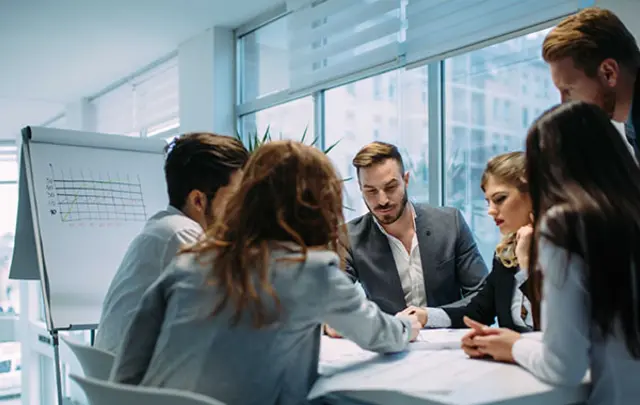 A group of professionals seated around a meeting table in a modern office, reviewing documents together, with a flip chart displaying a line graph in the background.