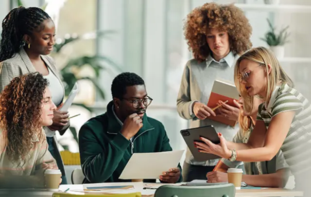 A diverse group of professionals collaborating around a table in a modern office, reviewing documents, a laptop, and a smartphone, with notebooks and coffee cups visible, illustrating teamwork, data analysis, and business decision-making.