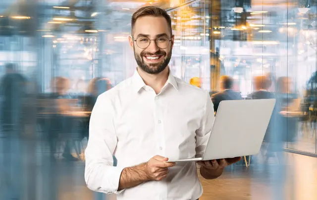 Person standing in a modern office holding a laptop, with a blurred busy workspace in the background.