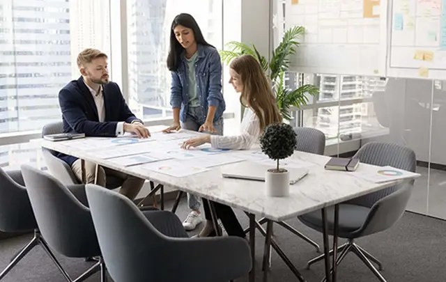 Three professionals collaborating around a table in a modern office, reviewing printed charts and financial data during a planning discussion