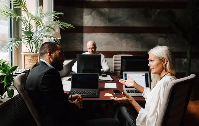 Three professionals sit around a meeting table with laptops, discussing work in a modern office. 