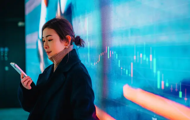 Person using a smartphone in front of a digital financial data display with charts and graphs, representing real-time analytics and FP&amp;A insights