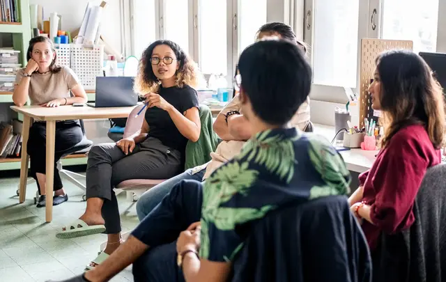 Team discussion in a bright office, with several people seated in a circle while one person speaks and others listen.